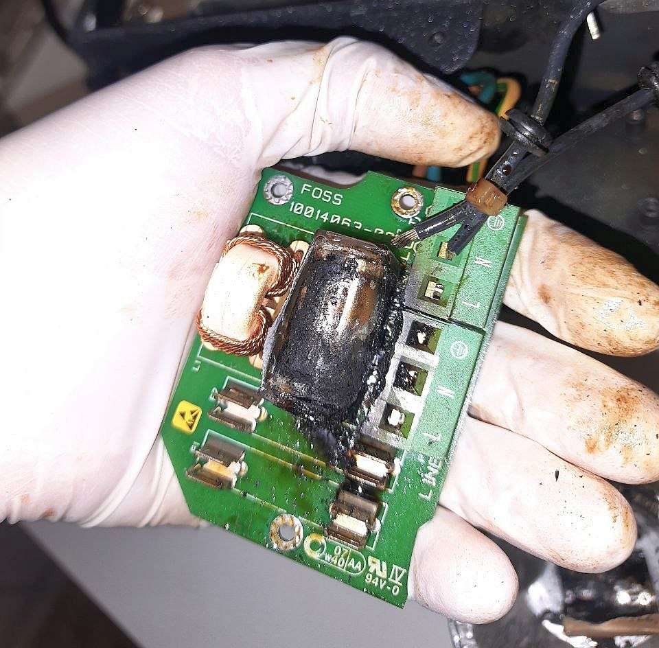 Mano con guante sosteniendo una placa de circuito impreso de un equipo FOSS con un condensador RIFA X2 completamente quemado, carbonizado y con restos de sustancia aceitosa.