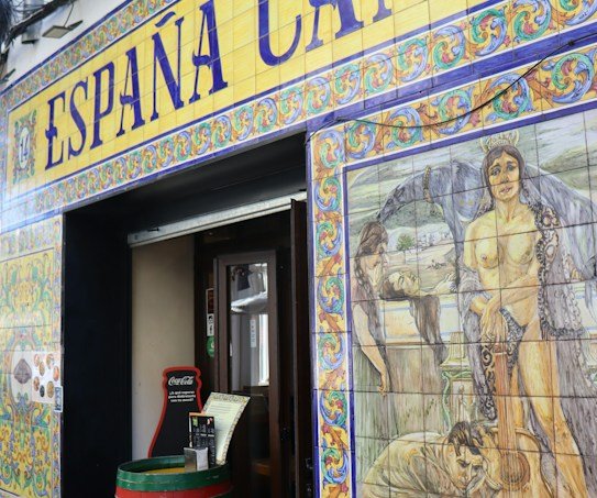 La entrada de un establecimiento español está decorada con azulejos de colores. Sobre la puerta, un rótulo grande dice «España Cañí», enmarcado por diseños ornamentales. A la derecha, un mural colorido representa escenas históricas o mitológicas, con personas y una guitarra. Junto a la entrada, un barril rojo sirve de mesa y aporta un aire rústico.